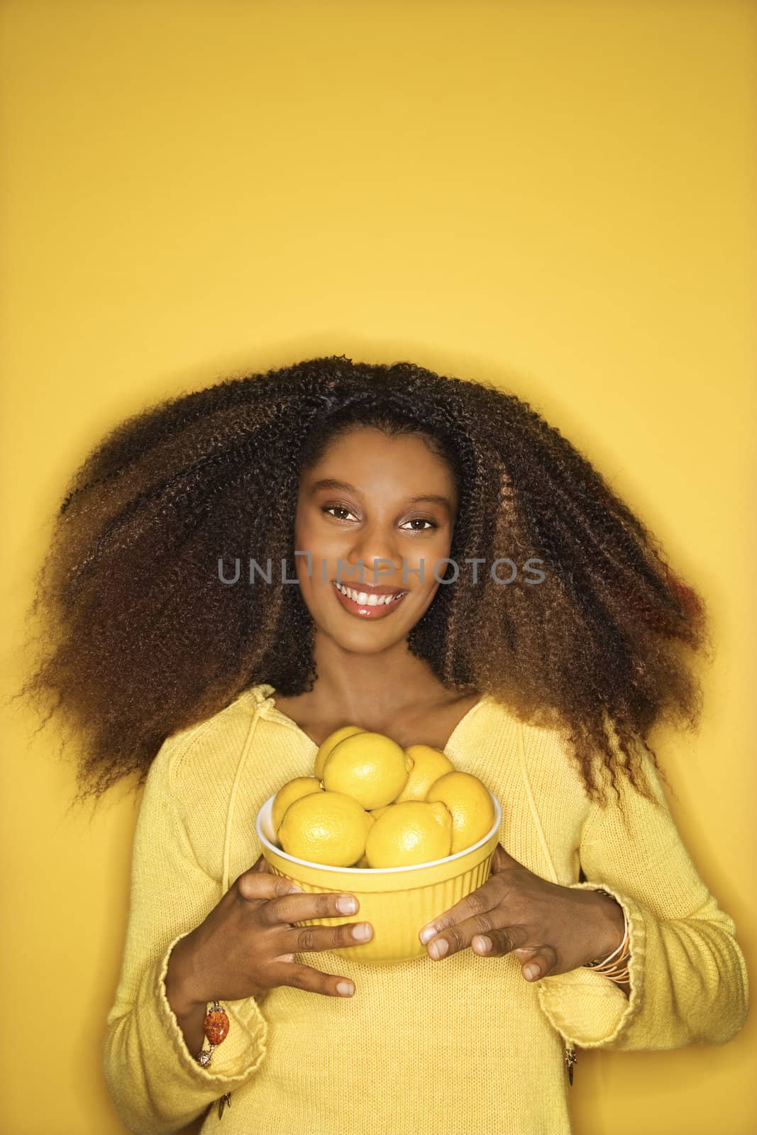 Portrait of young African-American adult woman smiling pleasantly against yellow background holding bowl of lemons.