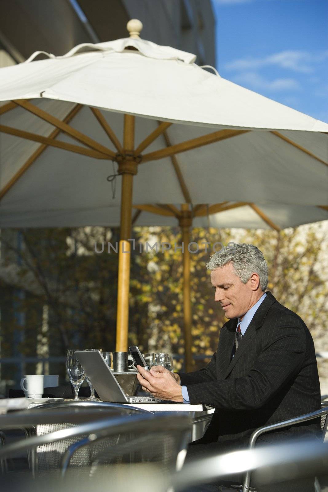 Prime adult Caucasian man in suit sitting at patio table outside with laptop and dialing cellphone.