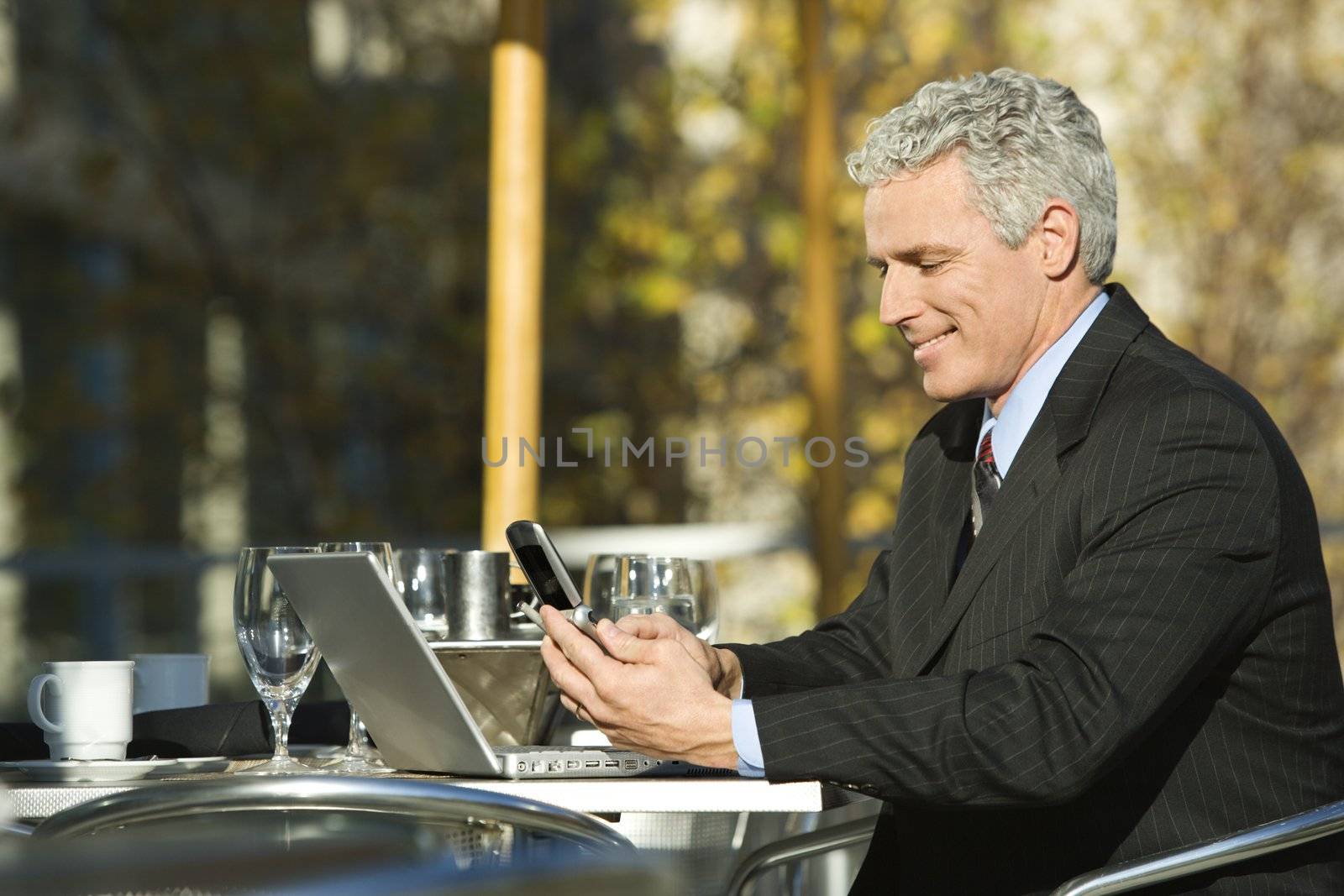 Profile of smiling prime adult Caucasian man in suit sitting at patio table outside with laptop and dialing cellphone.