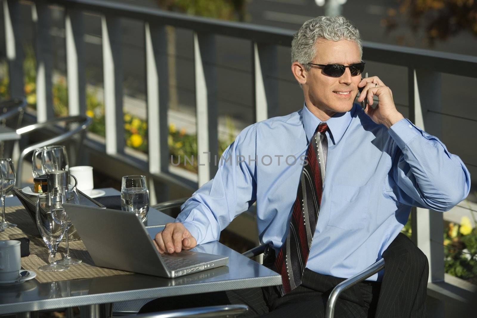 Prime adult Caucasian man in suit sitting at patio table ouside with laptop and talking on cellphone.