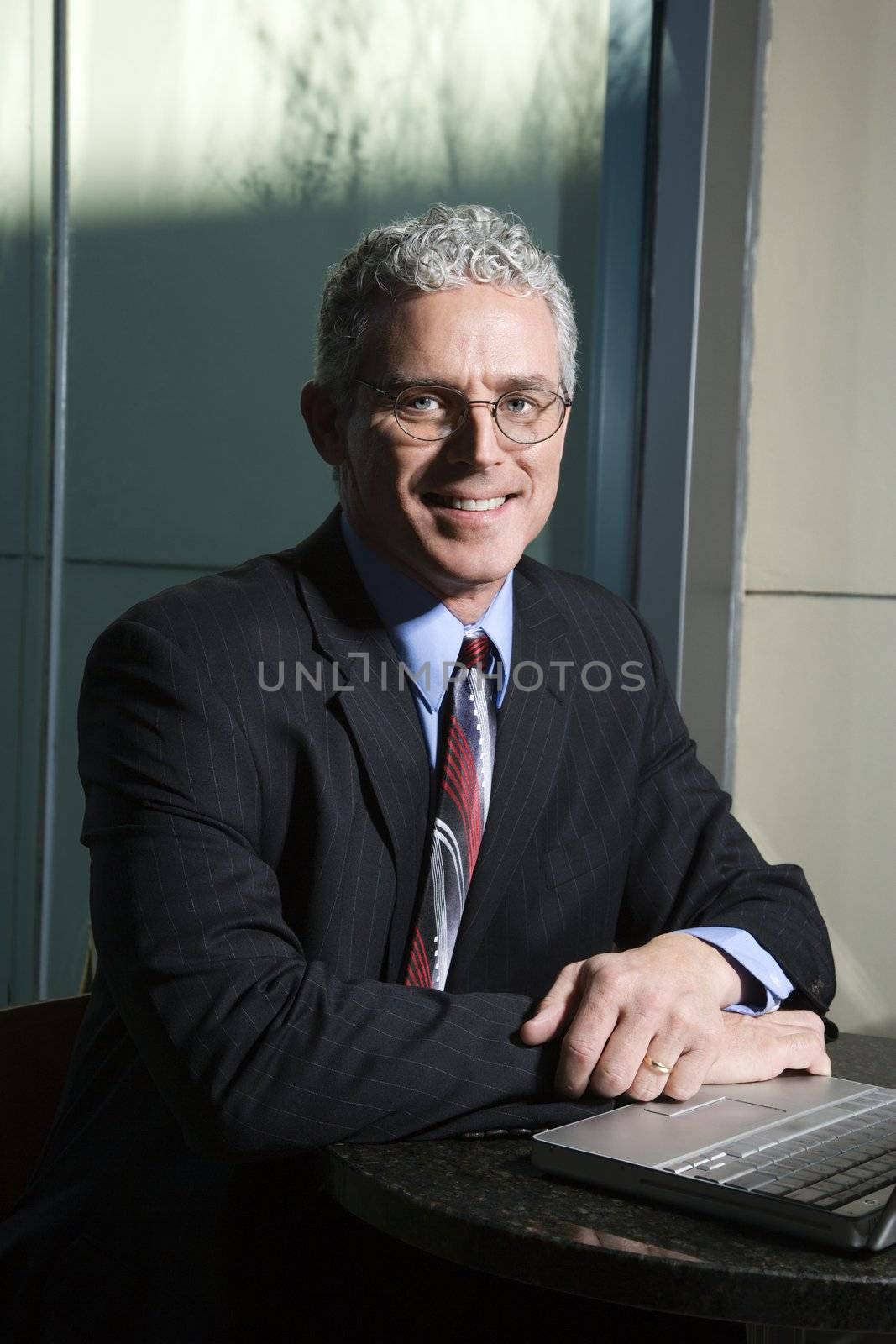 Close up of prime adult Caucasian man in suit sitting at table with laptop looking at viewer and smiling.