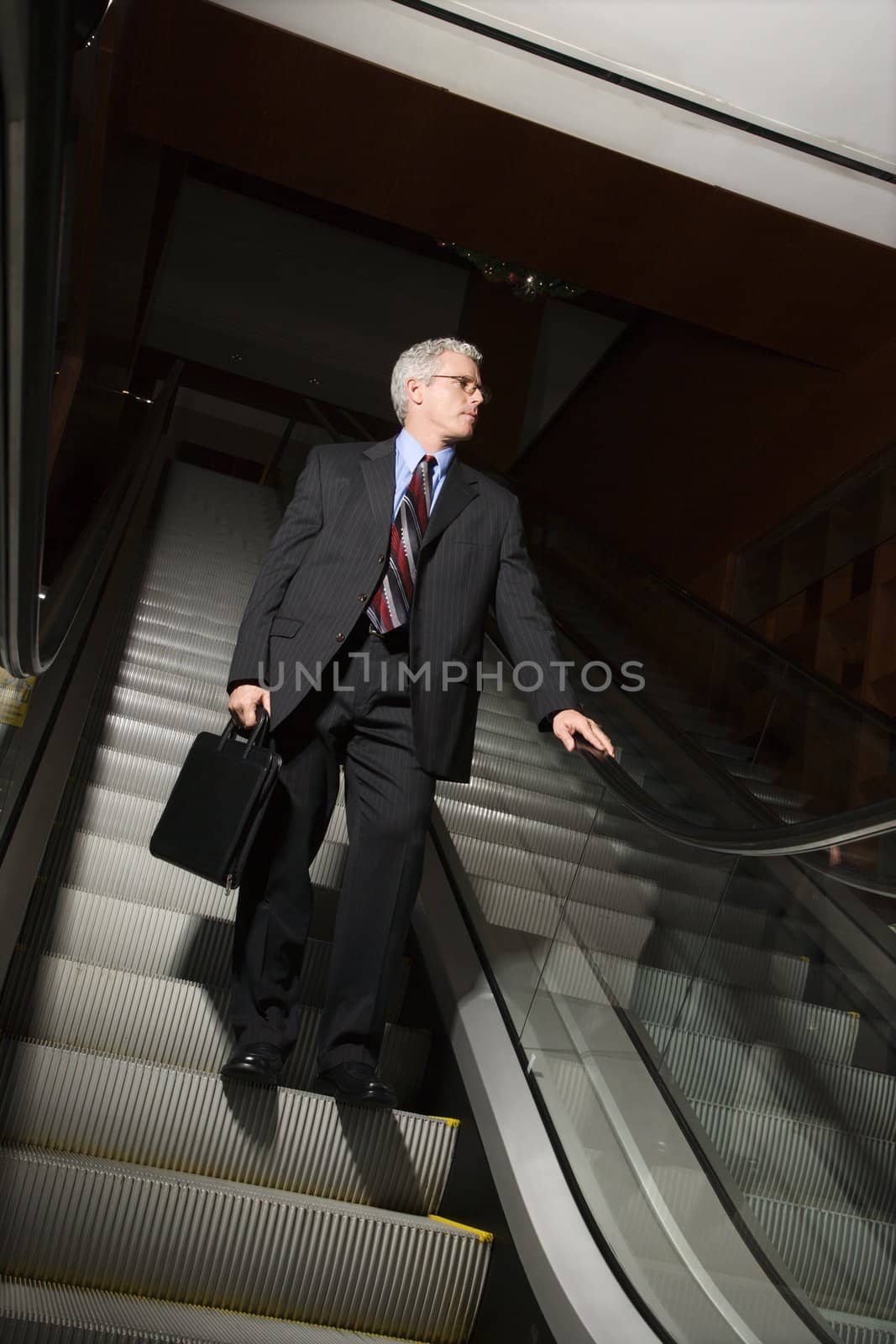 Prime adult Caucasian man in suit holding briefcase standing on down escalator holding rail.