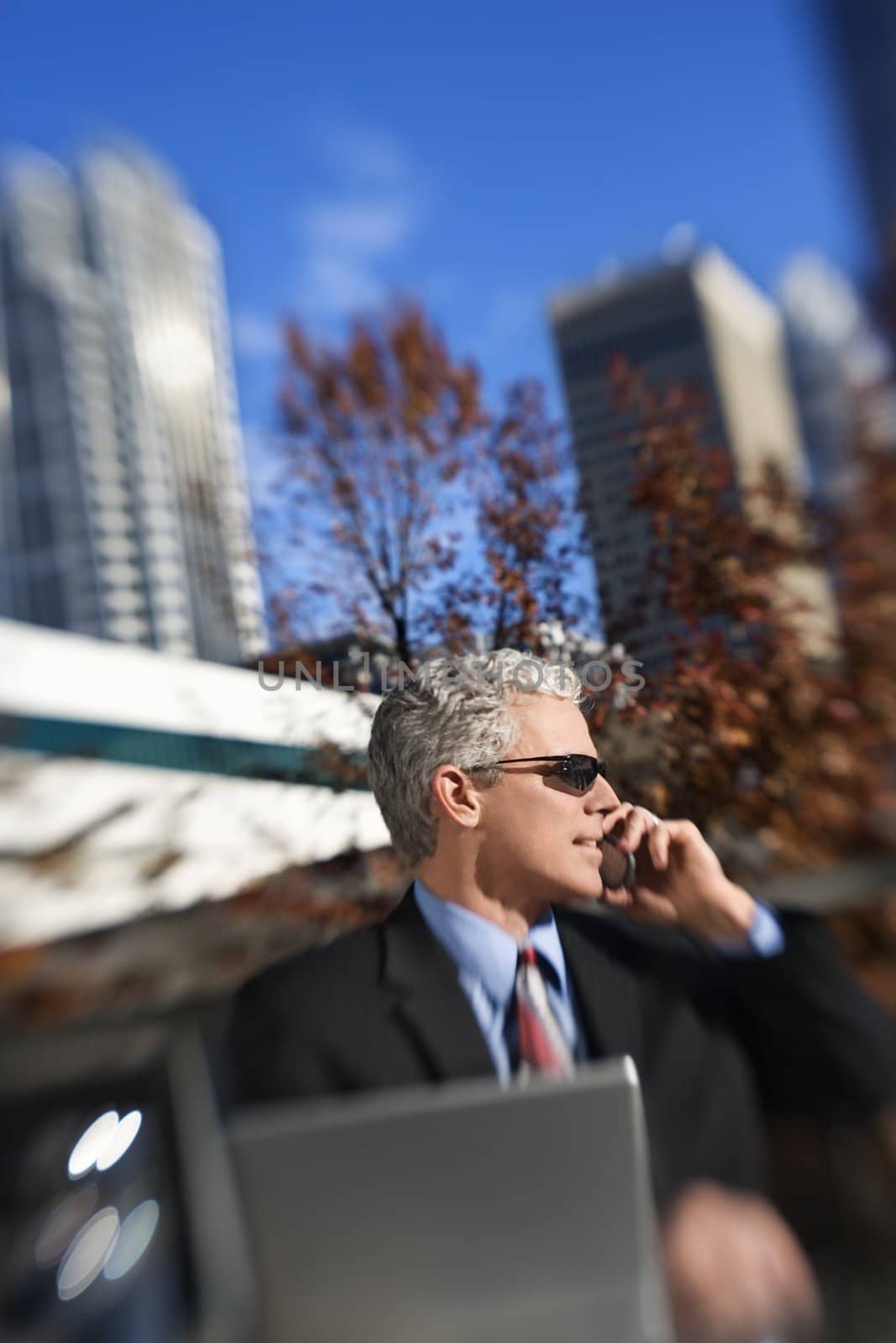 Prime adult Caucasian man in suit sitting at patio table outside with laptop talking on cellphone wearing sunglasses with buildings in background.
