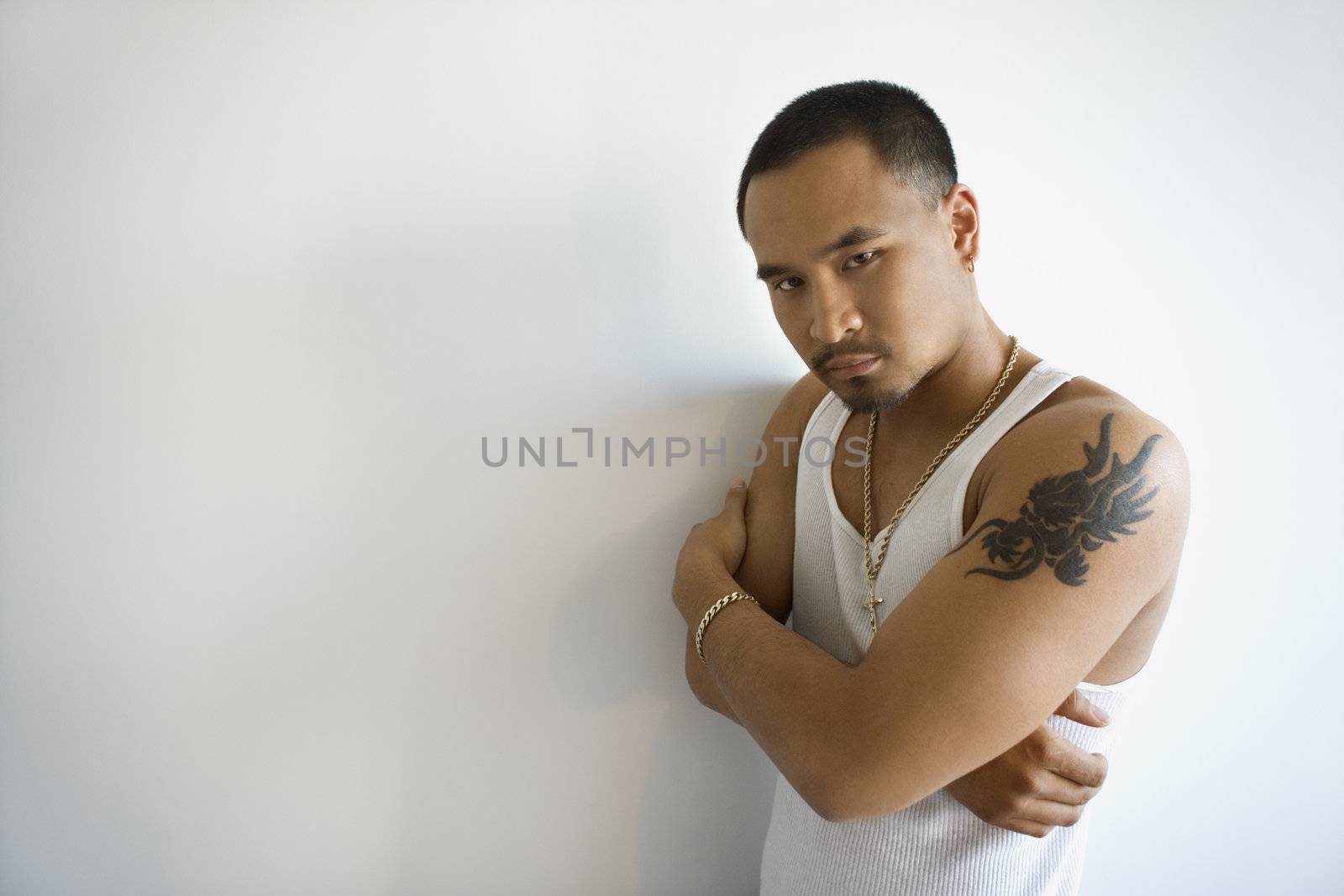 Portrait of Asian young adult man leaning against white wall with arms crossed looking at viewer.
