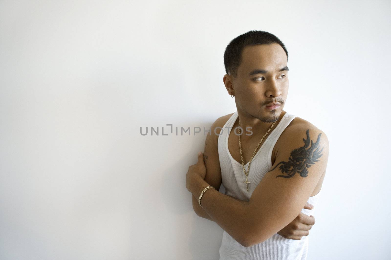 Portrait of Asian young adult man leaning against white wall with arms crossed looking over shoulder.