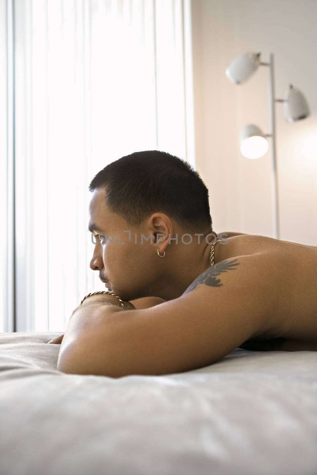 Profile of shirtless Asian young adult man lying on bed resting head on arms looking out window.