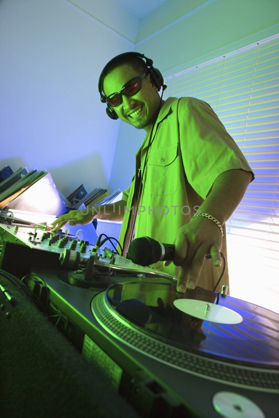 Asian young adult male DJ wearing sunglasses behind mixing equipment with hand on record turntable looking at viewer smiling.