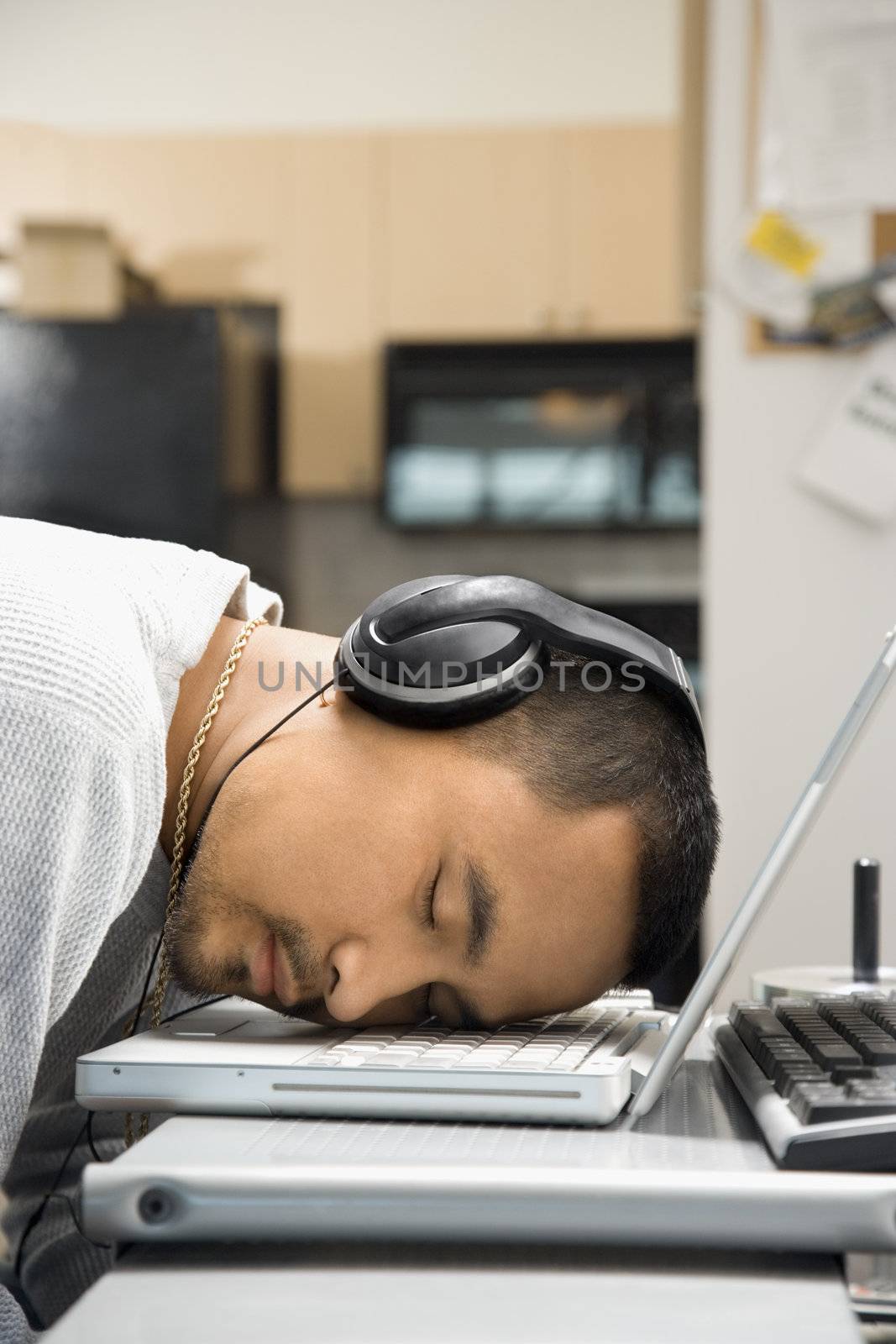 Close-up of Asian young adult man sleeping with head on laptop keyboard and wearing headphones.