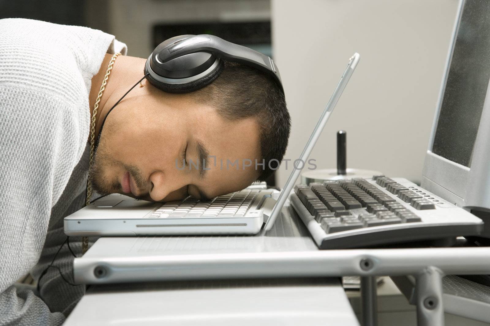Close-up of Asian young adult man sleeping with head on laptop keyboard and wearing headphones.