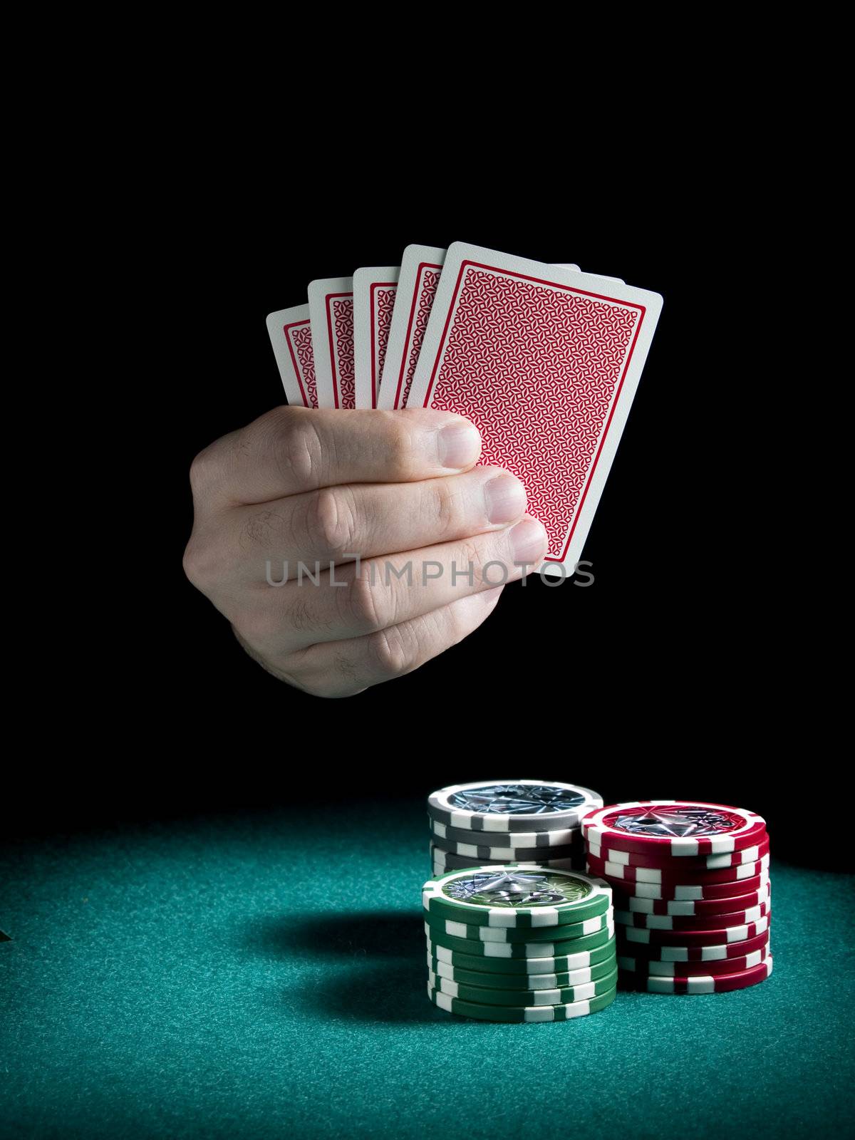 A man's hand holding four cards over three piles of different colors chips on a green felt.