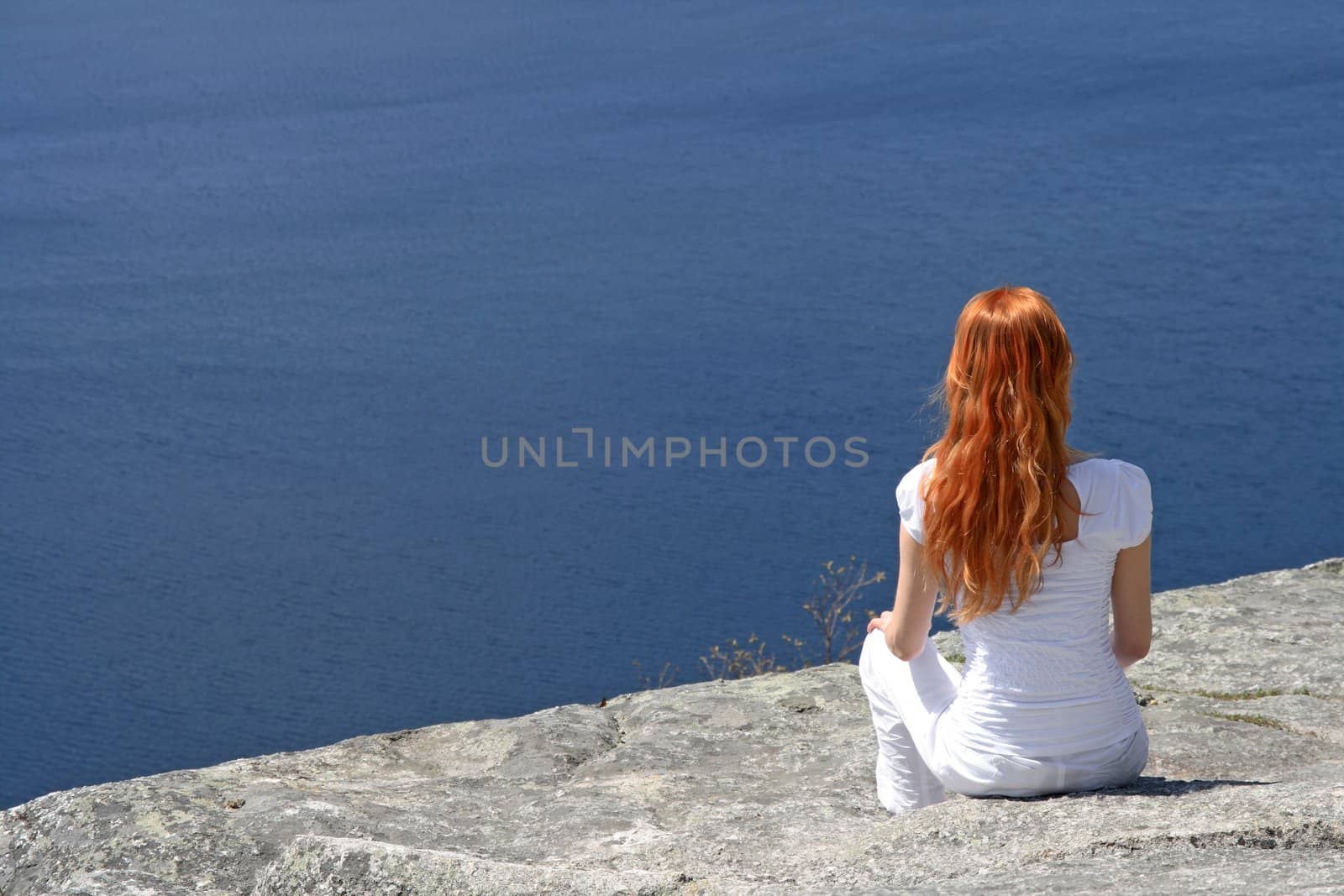 Red-haired girl sitting on a rock and looking over blue water.