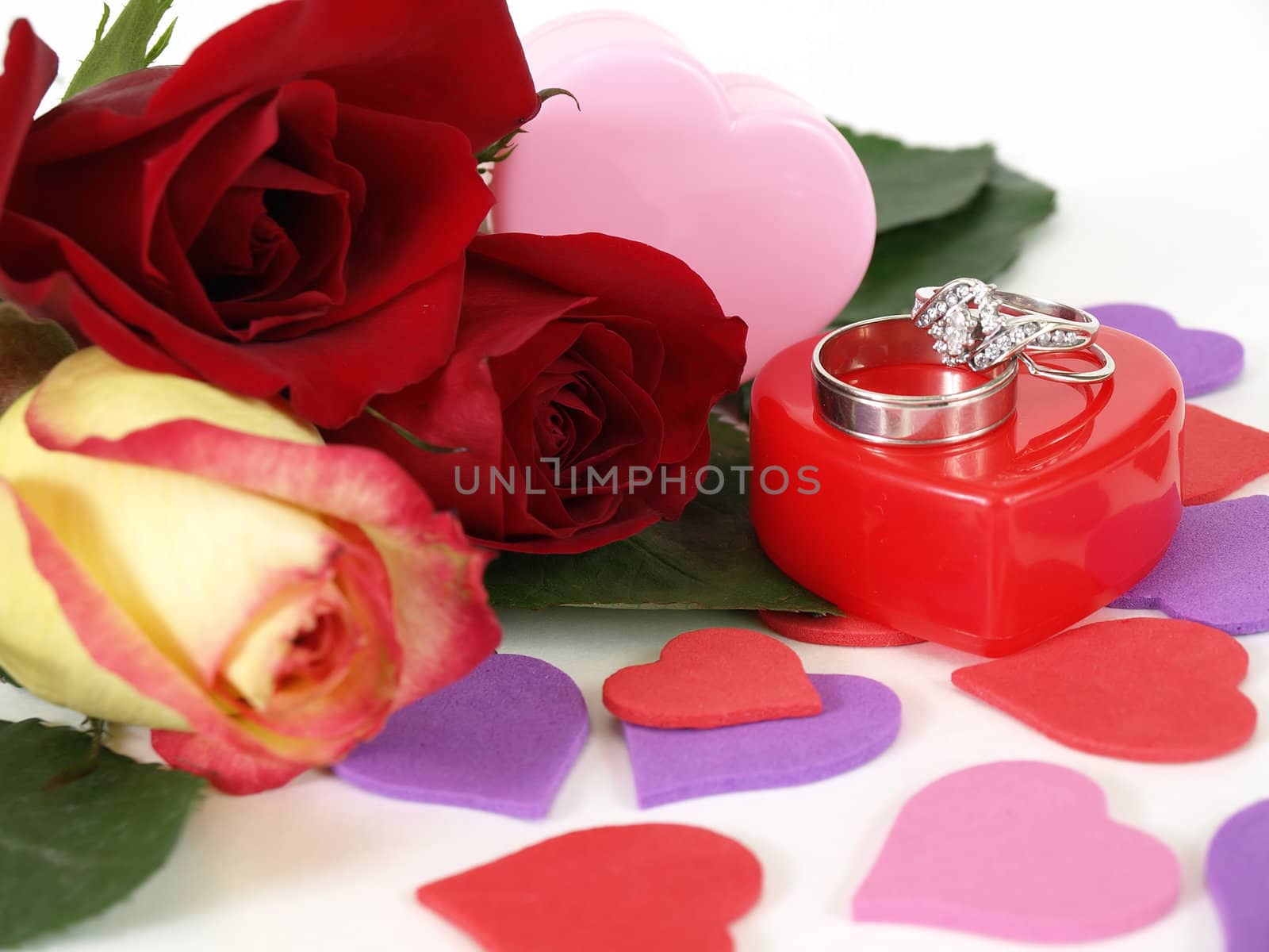 Wedding rings sit atop a red plastic box. Three roses lay next to it, various heart shapes scattered around.