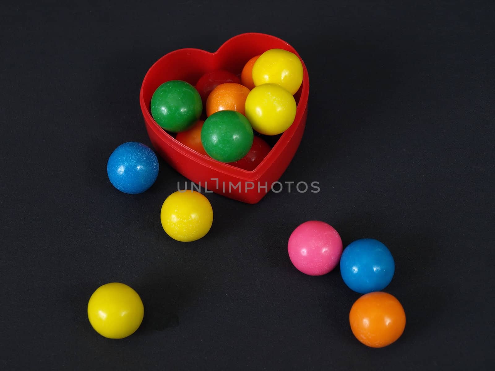 A small heart shaped container with colored gumballs, over a black background.