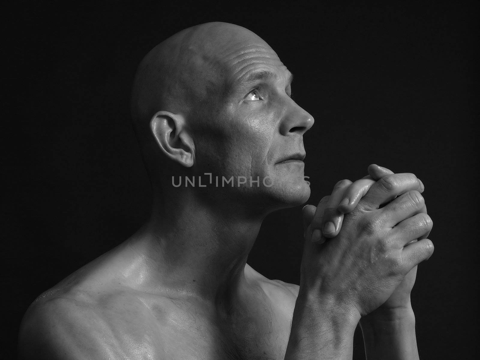 A bald shirtless man clasps his hands in prayer, eyes pleading skyward, over a black background. In black and white.