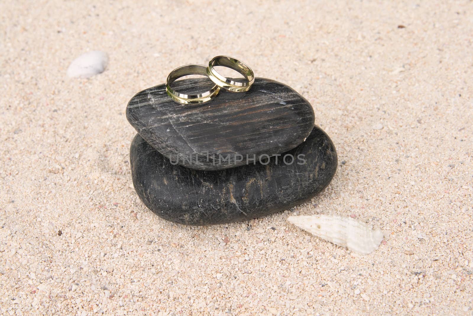 A pair of gold wedding rings placed upon black pebbles on the sand beside a white seashell