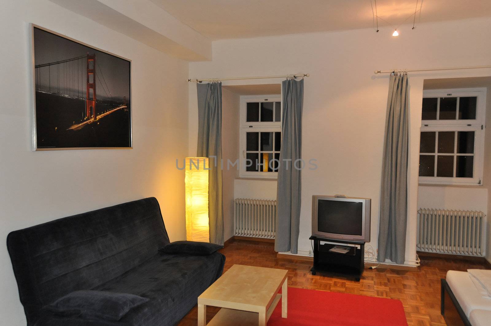 View of a large and modern hotel room with sofa, table, tv set, carpet, lamp and windows