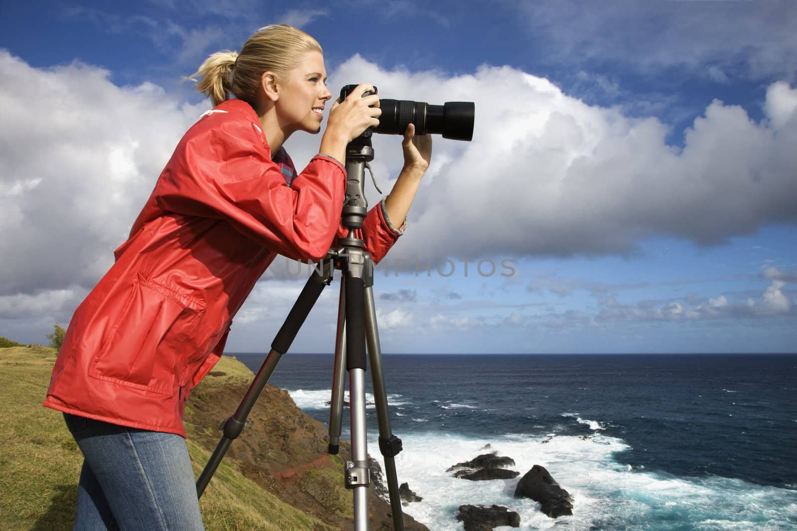 Caucasian mid-adult woman looking through camera on tripod on cliff overlooking ocean in Maui, Hawaii.