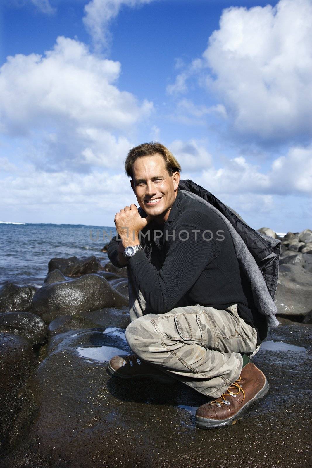 Caucasian mid-adult man kneeling on rocky coast looking at viewer smiling in Maui, Hawaii.