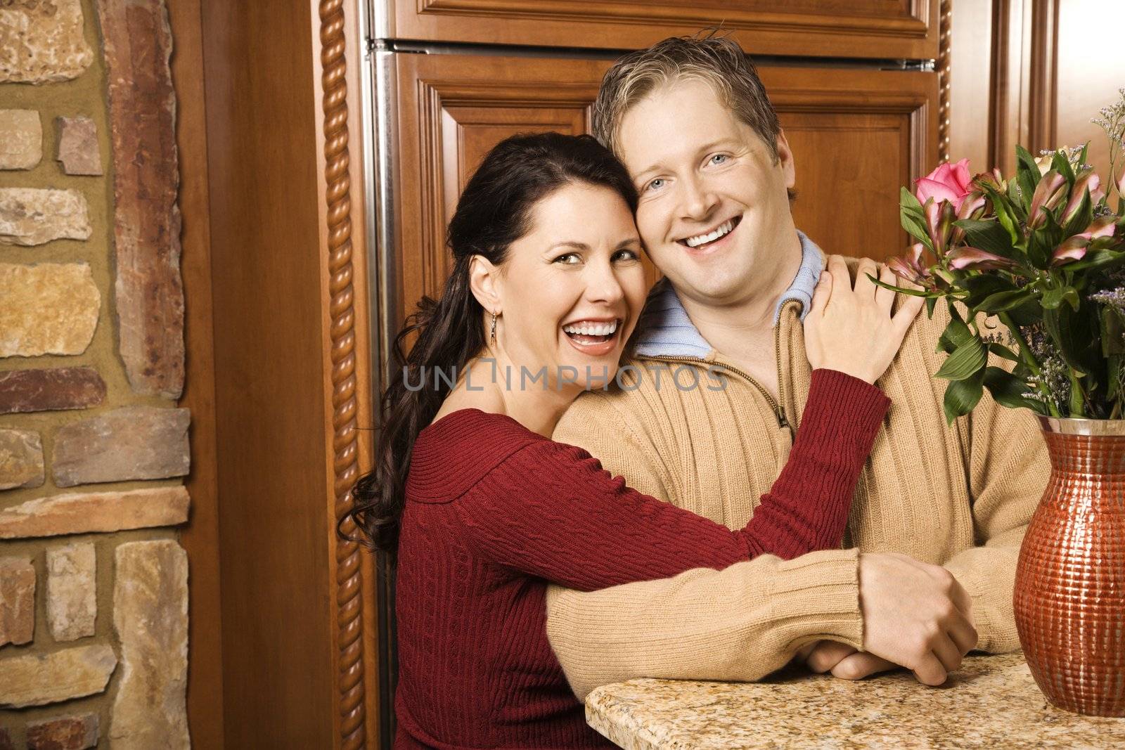 Caucasian woman with arms around Caucasian man in kitchen smiling and looking at viewer.