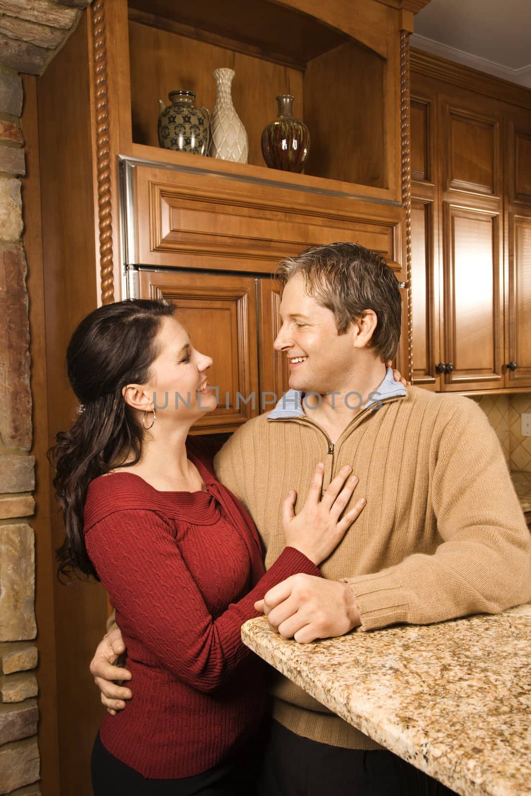 Caucasian woman and Caucasian man with arms around each other leaning on kitchen counter and smiling at each other.