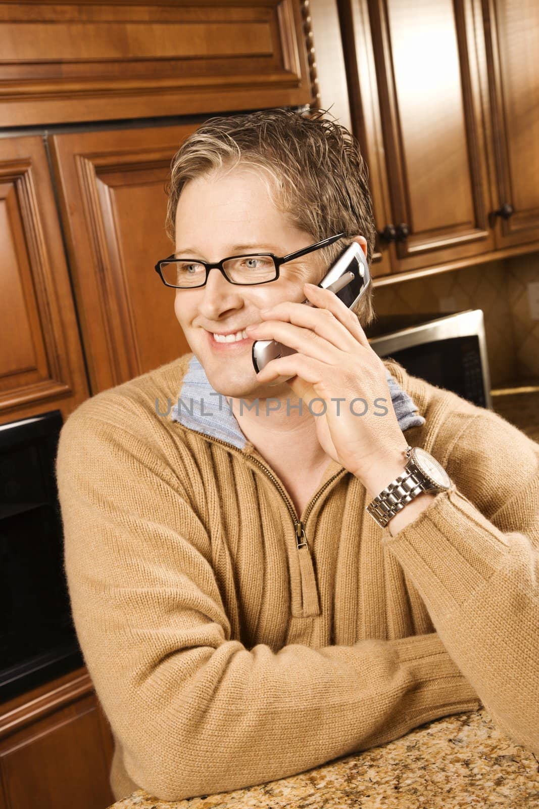 Caucasian man smiling and talking on cellphone while leaning on kitchen counter.
