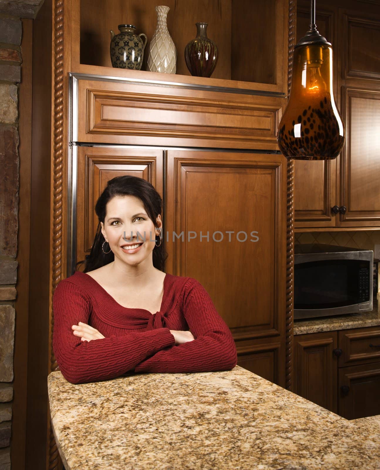 Caucasian woman leaning on marble kitchen counter smiling with arms crossed and looking at viewer.