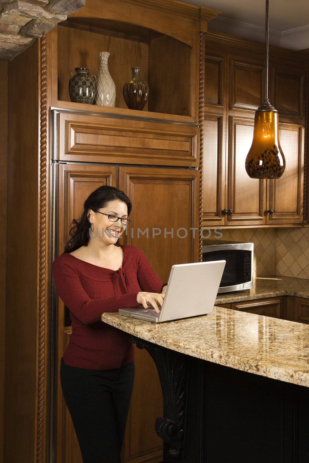 Caucasian woman standing at kitchen counter typing on laptop computer and smiling.