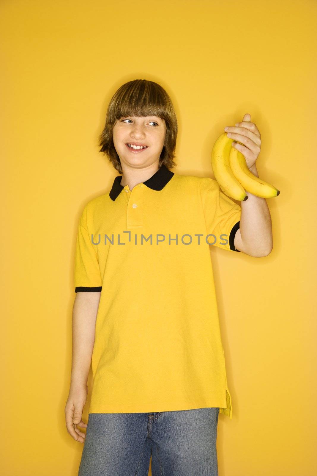 Portrait of smiling Caucasian boy holding bunch of bananas standing against yellow background.