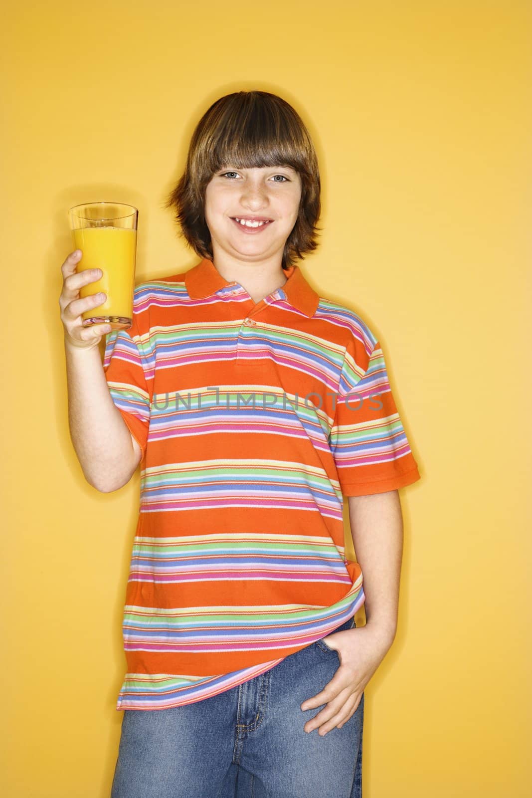 Portrait of smiling Caucasian boy holding glass of orange juice and standing against yellow background.