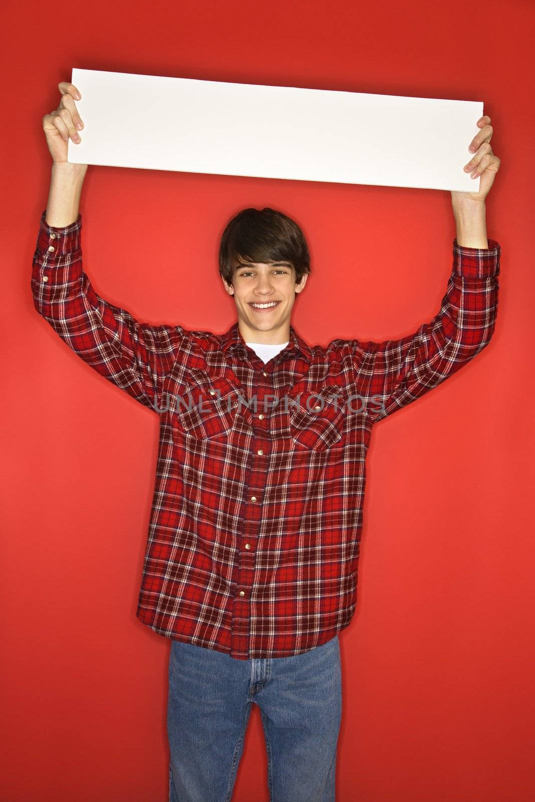 Portrait of Caucasian teen boy holding blank sign above his head standing against red background.