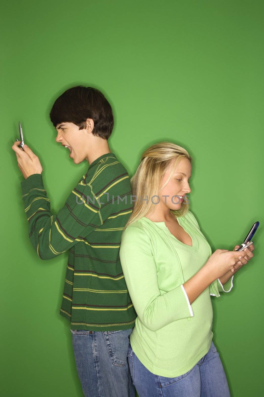 Portrait of Caucasian teen boy and girl on cellphones standing with backs to eachother against green background.