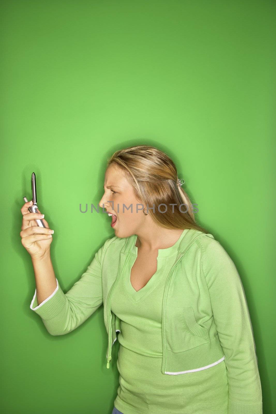 Portrait of blond Caucasian teen girl looking at cellphone with shock and disbelief against green background.