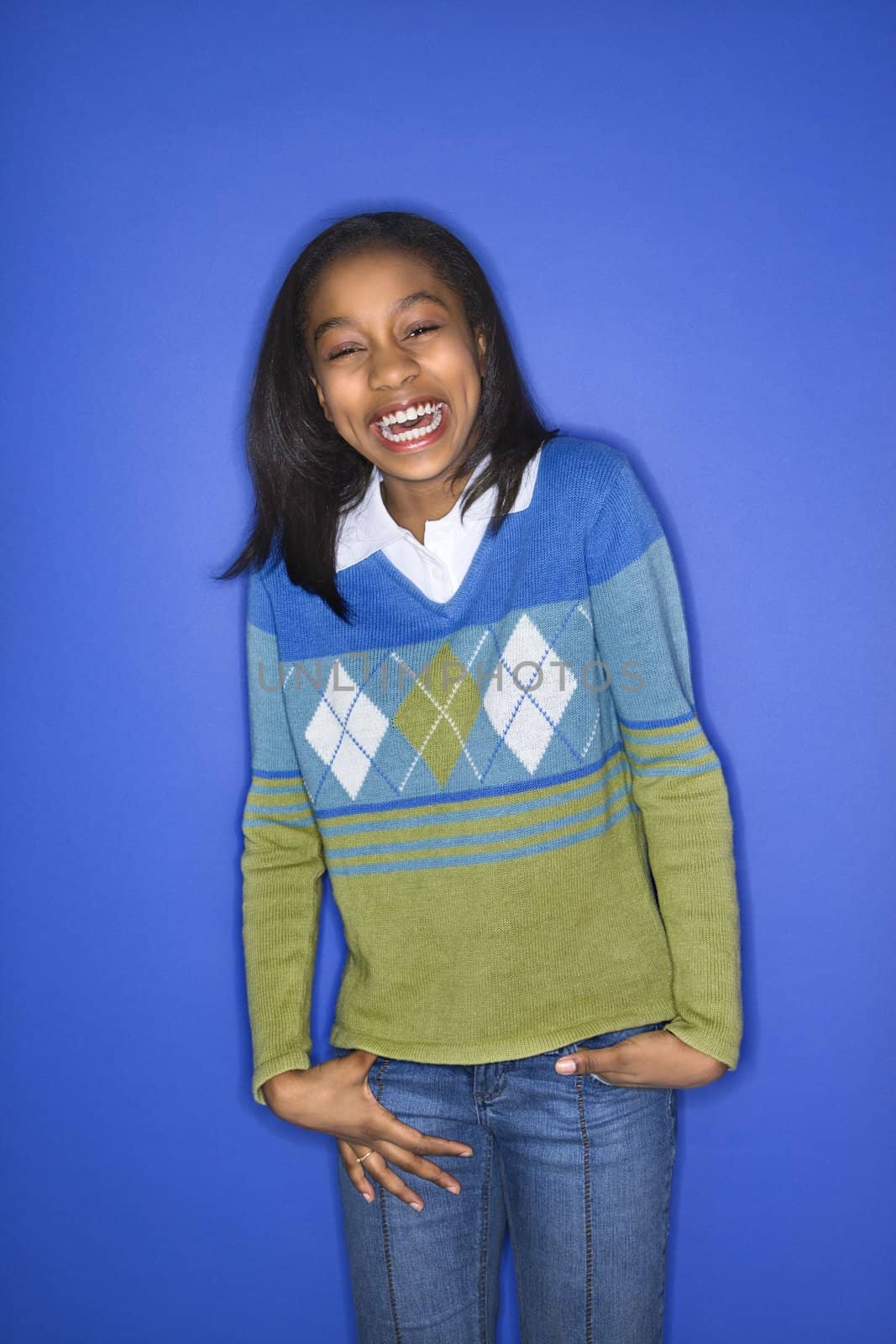 Portrait of African-American girl with hands in her pockets smiling standing against blue background.