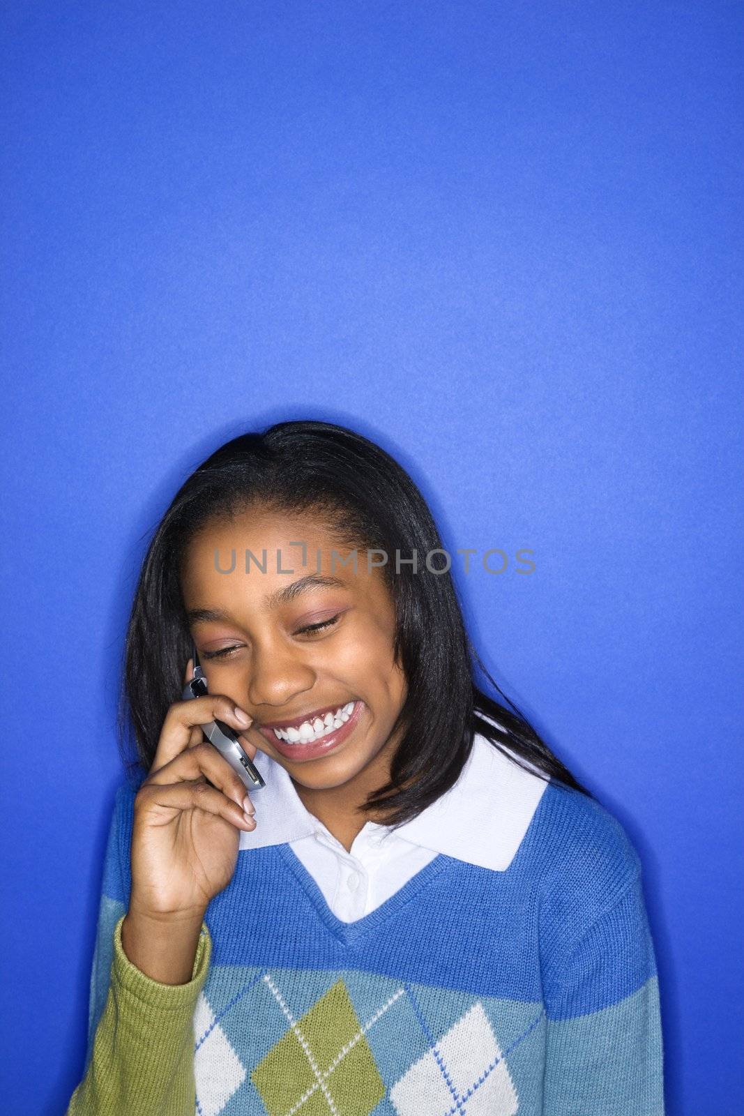 Portrait of African-American teen girl talking on cellphone smiling standing in front of blue background.