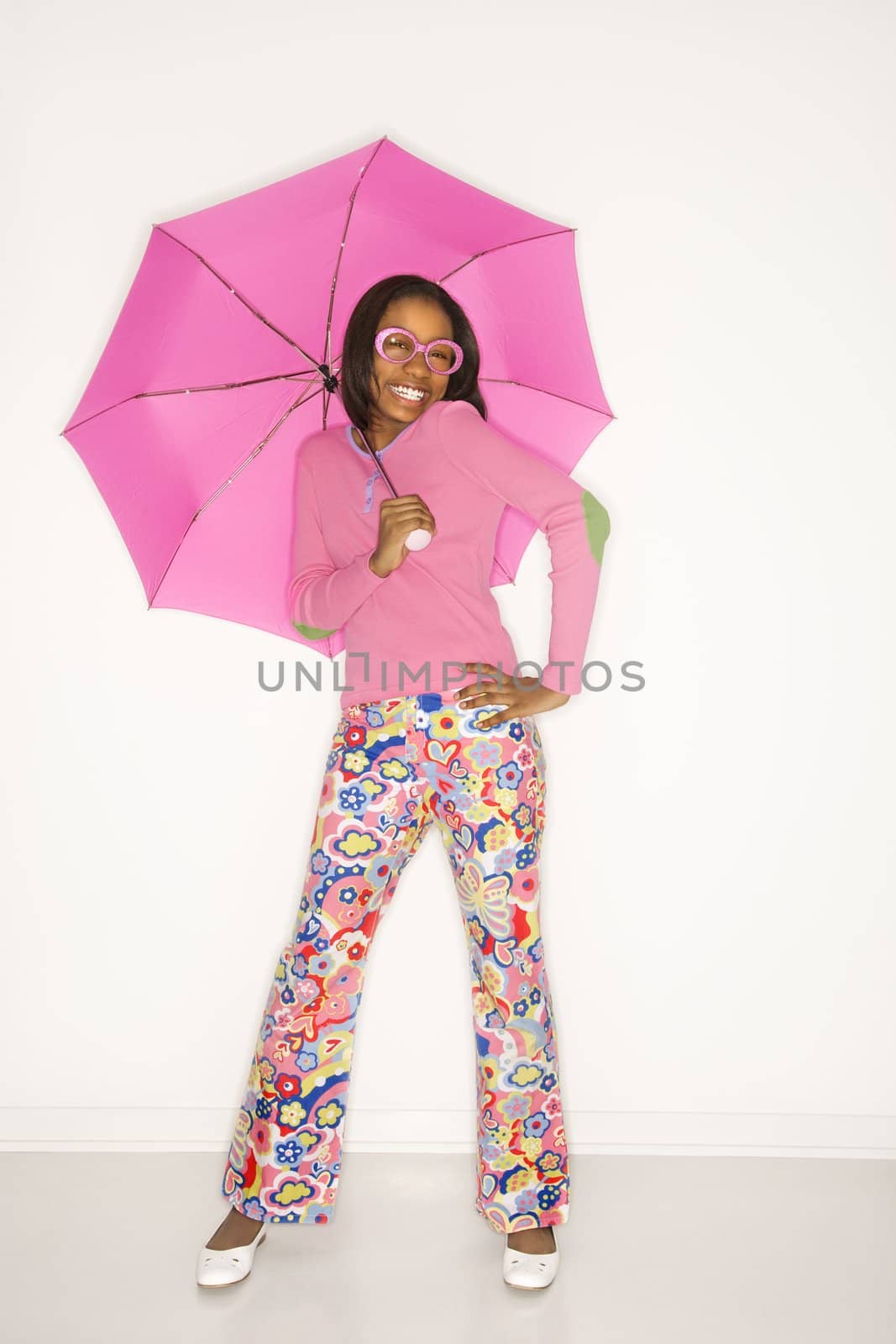 Portrait of smiling African-American teen girl holding a pink umbrella with hand on hip standing against white background.