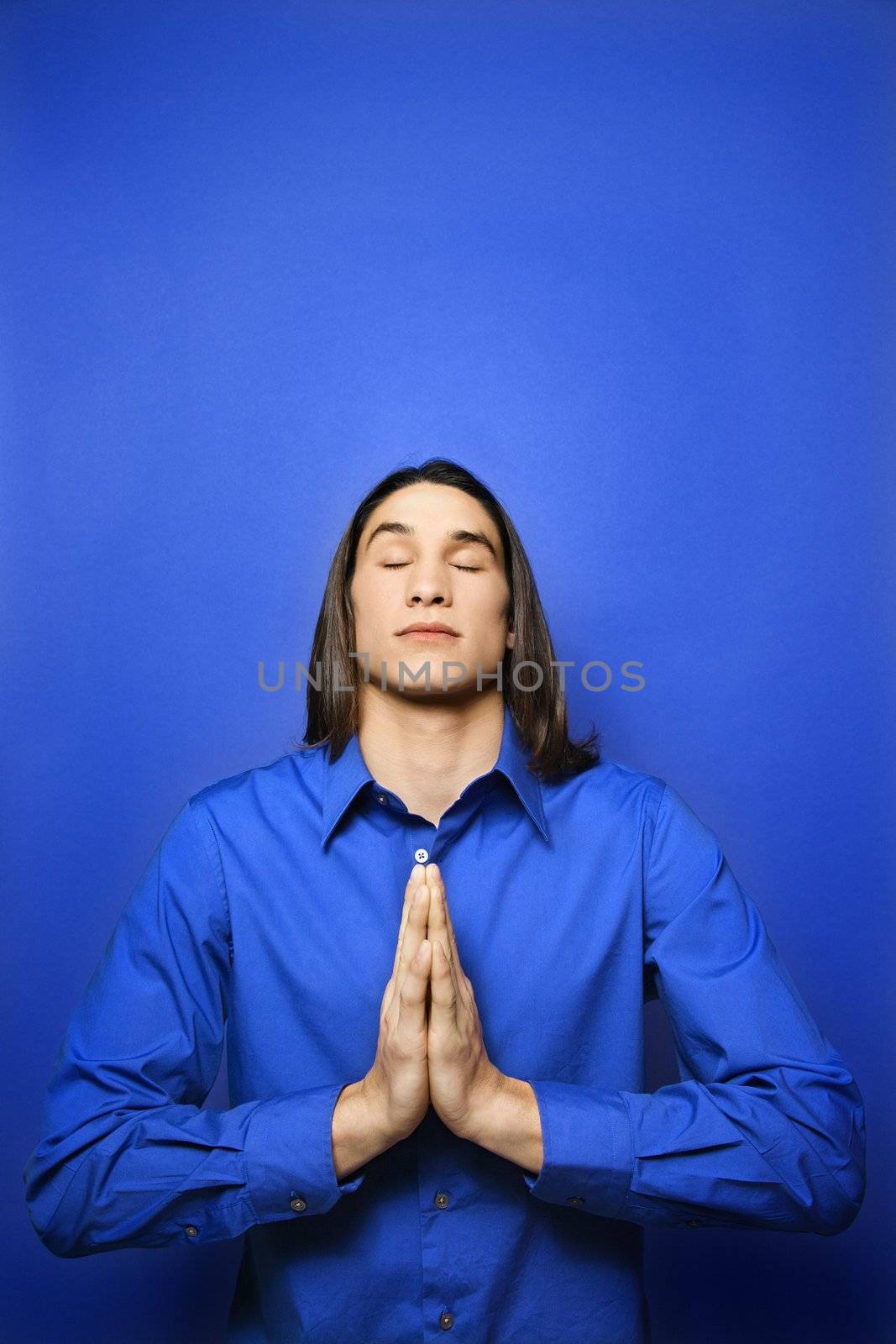 Portrait of Asian-American teen boy with palms pressed together in prayer position standing against blue background.
