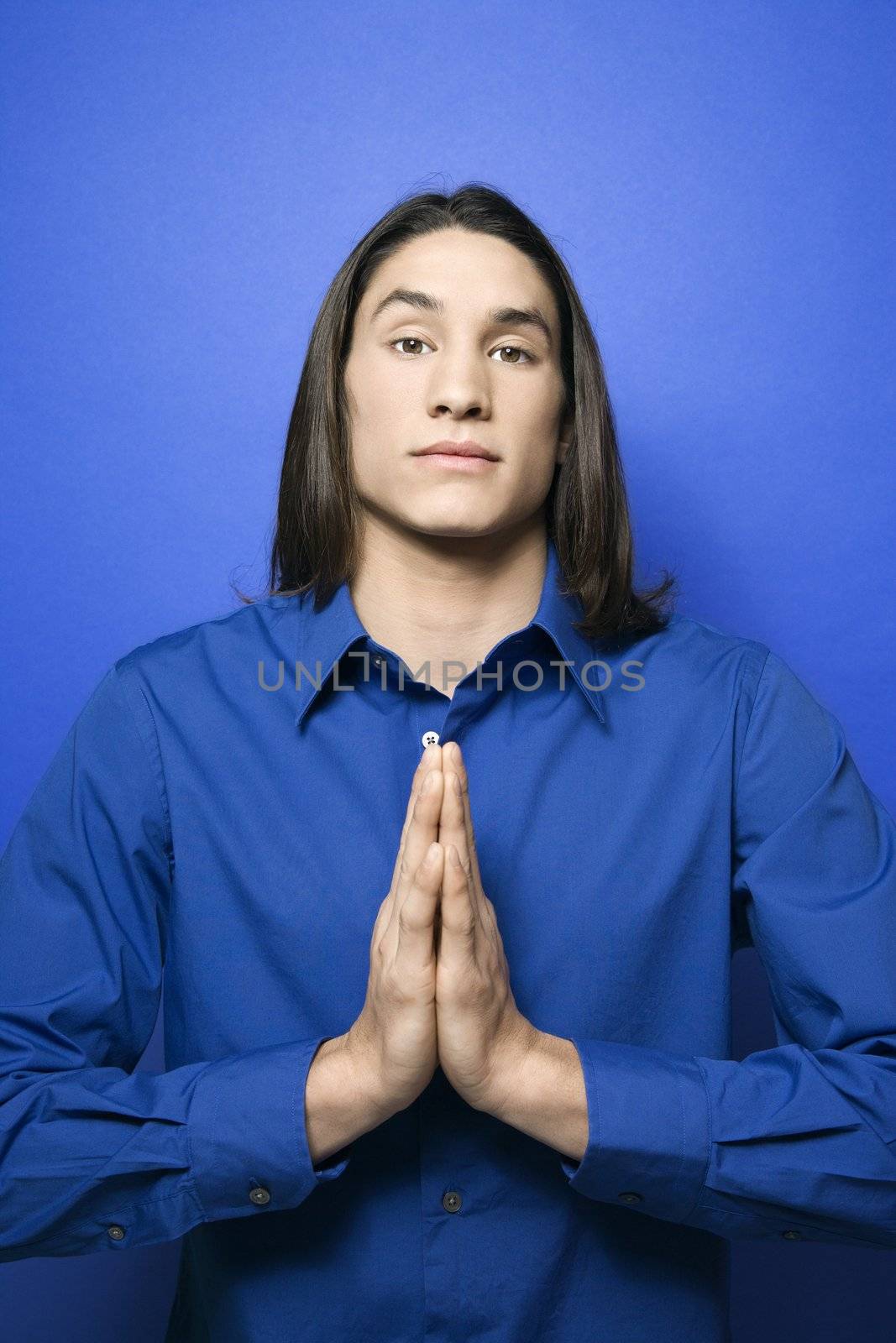 Portrait of Asian-American teen boy with hands pressed together in prayer position standing against blue background.