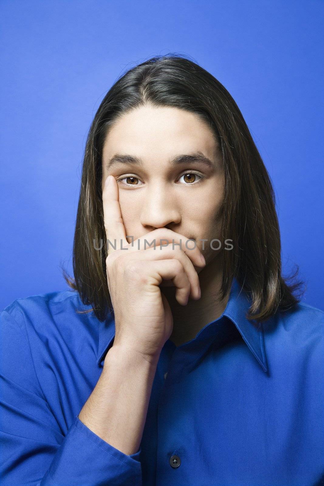 Portrait of Asian-American teen boy with hand covering chin and finger on side of face against blue background.
