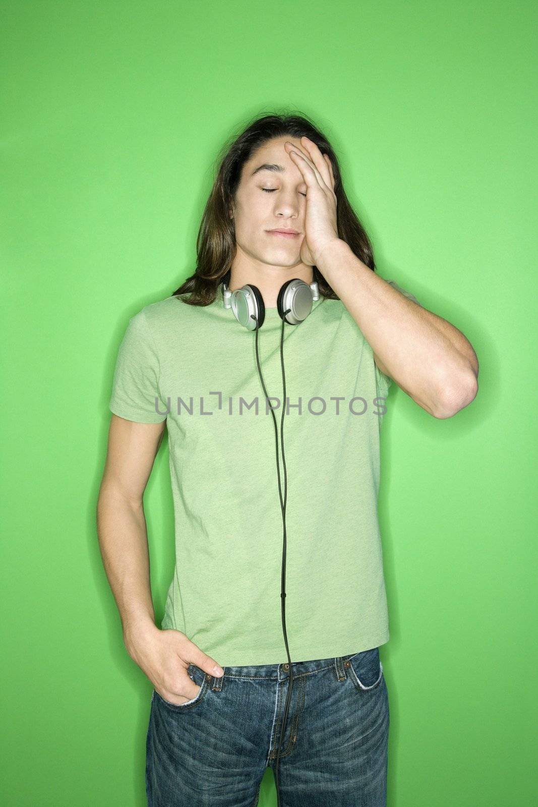 Portrait of Asian-American teen boy with headphones around neck and hand on forehead standing against green background.