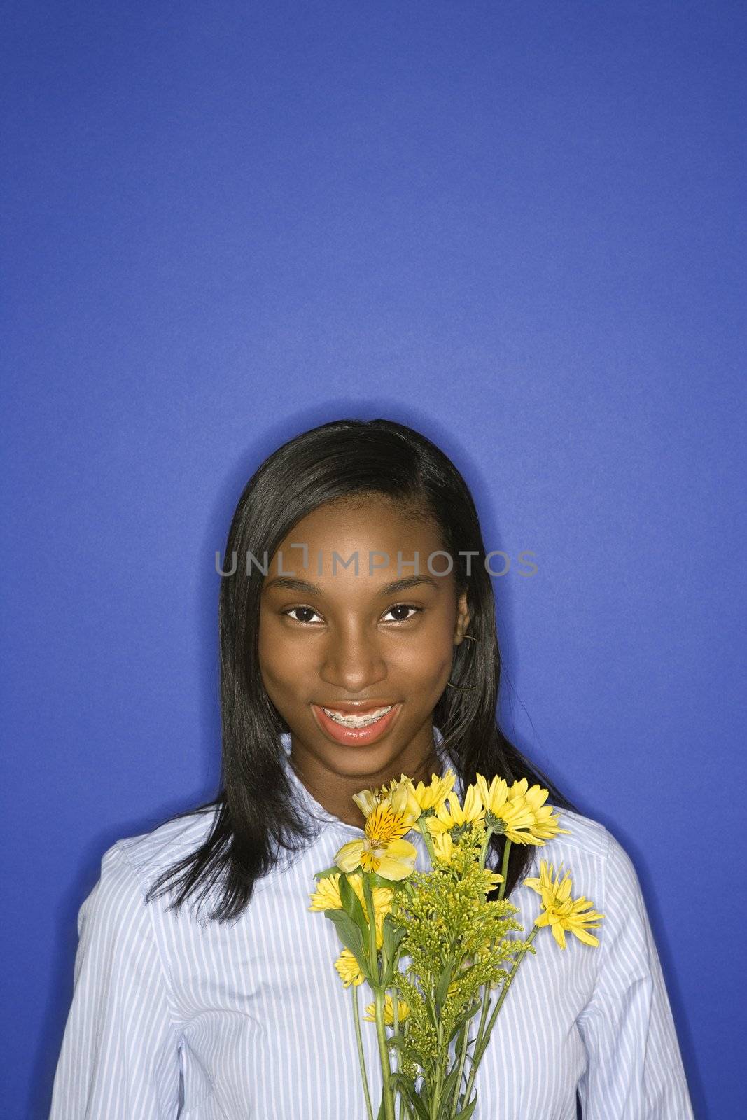 Portrait of smiling African-American teen girl with bouquet of yellow flowers in front of blue background.
