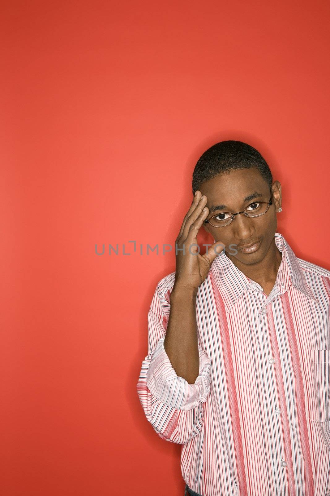 Portrait of African-American teen boy with glasses and hand on forehead against red background.