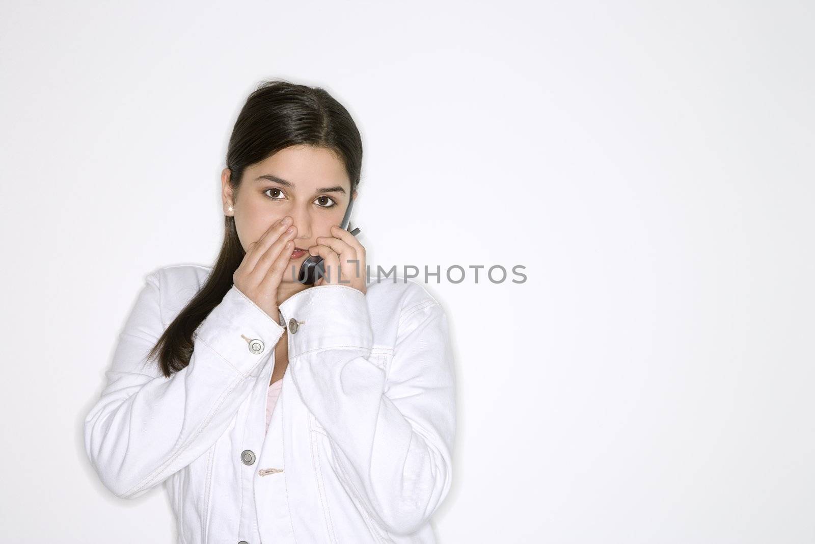 Portrait of Caucasian teen girl whispering into cellphone standing against white background.