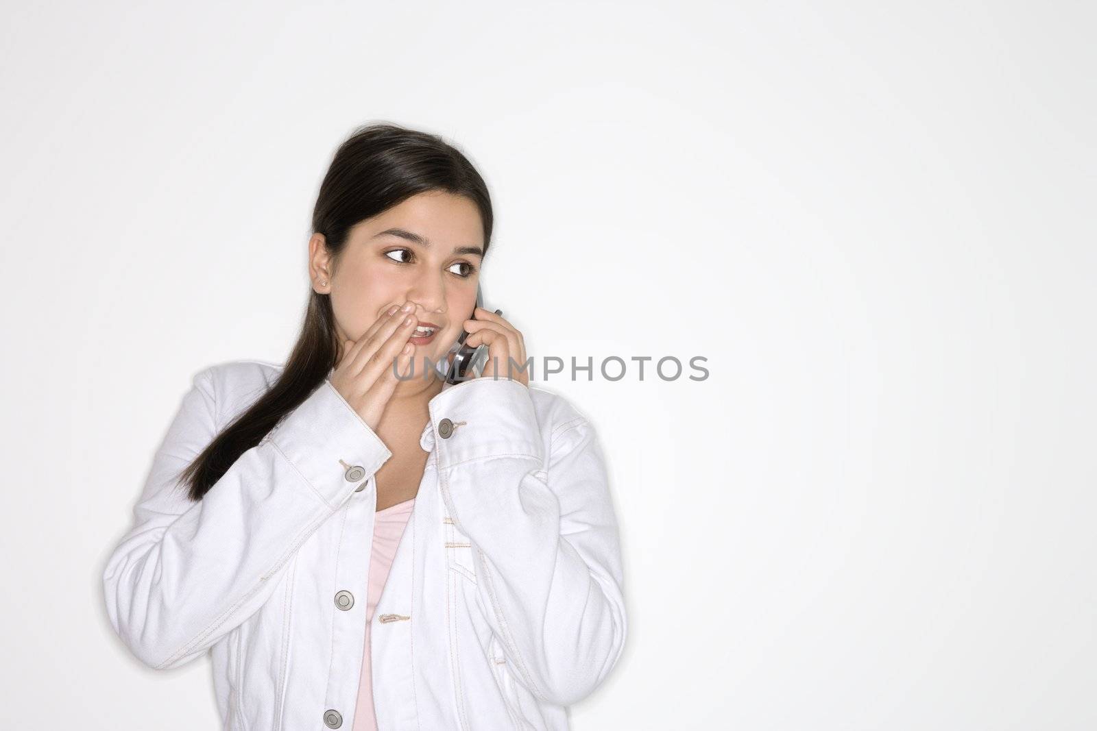 Portrait of Caucasian teen girl whispering into cellphone standing against white background.