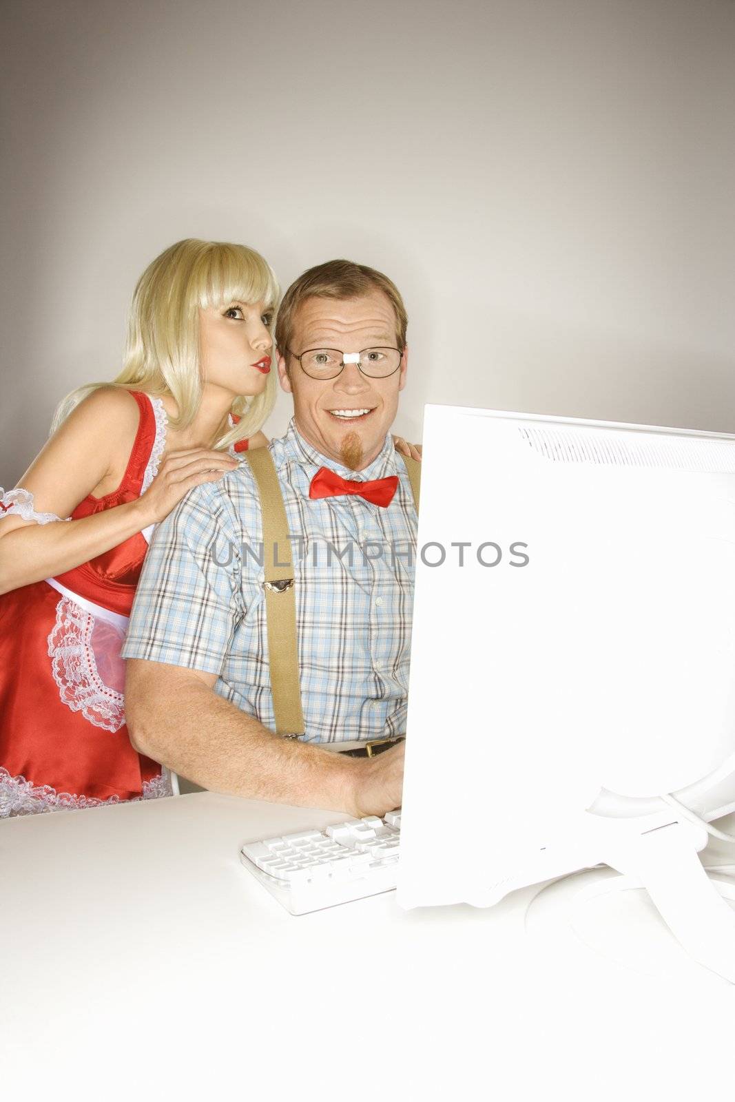Caucasian young blonde woman dressed in french maid outfit whispering to Caucasian young man sitting behind computer dressed like nerd.