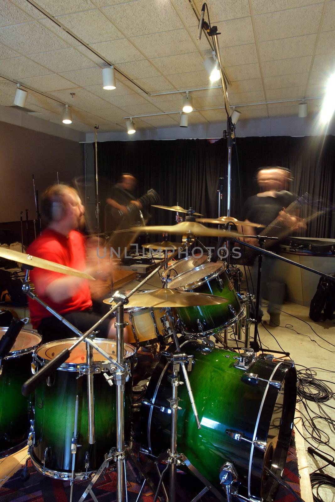 A bass player, guitar player and drummer recording tracks in a recording studio.  Slow shutter speed with ambient light - players have motion blur from slow shutter speed.
