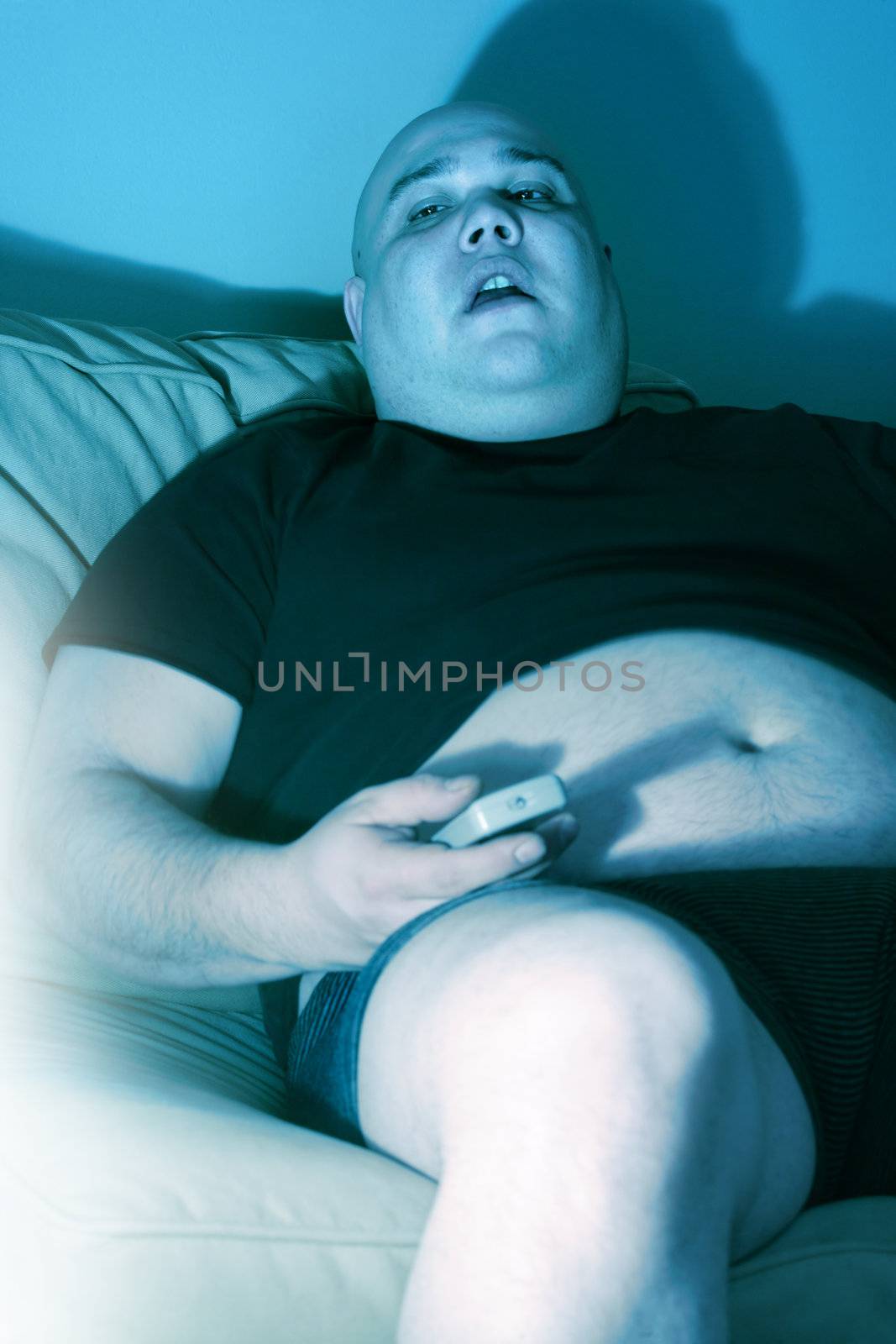 Lazy overweight male sitting on a couch watching television.  Harsh blue lighting from television with slow shutter speed to create TV watching atmosphere. Selective focus on the eyes.
