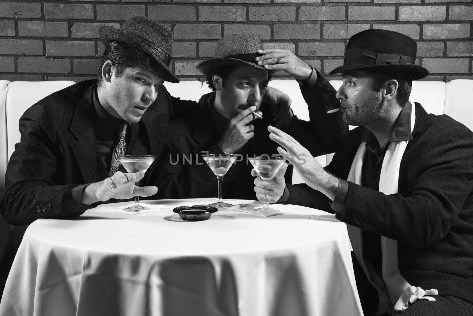 Three Caucasian prime adult males in retro suits sitting at table drinking and smoking and talking.
