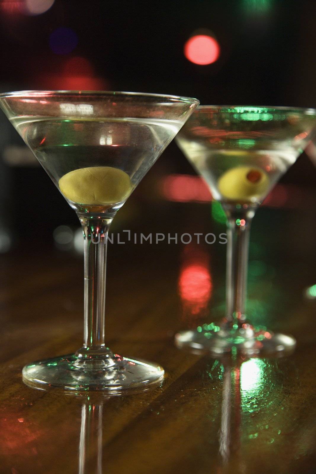 Two martinis with olives on bar with red and green lights in background.