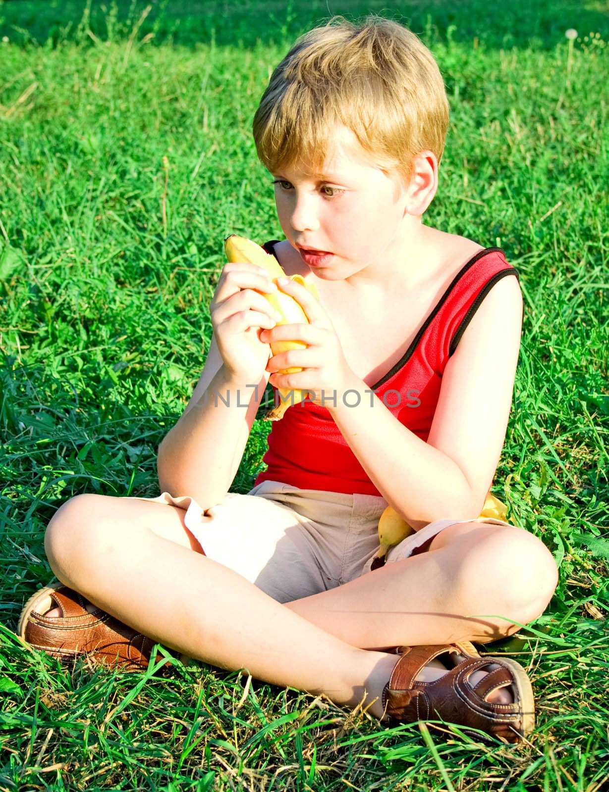 The little boy sits on a green grass and eats a ripe yellow banana