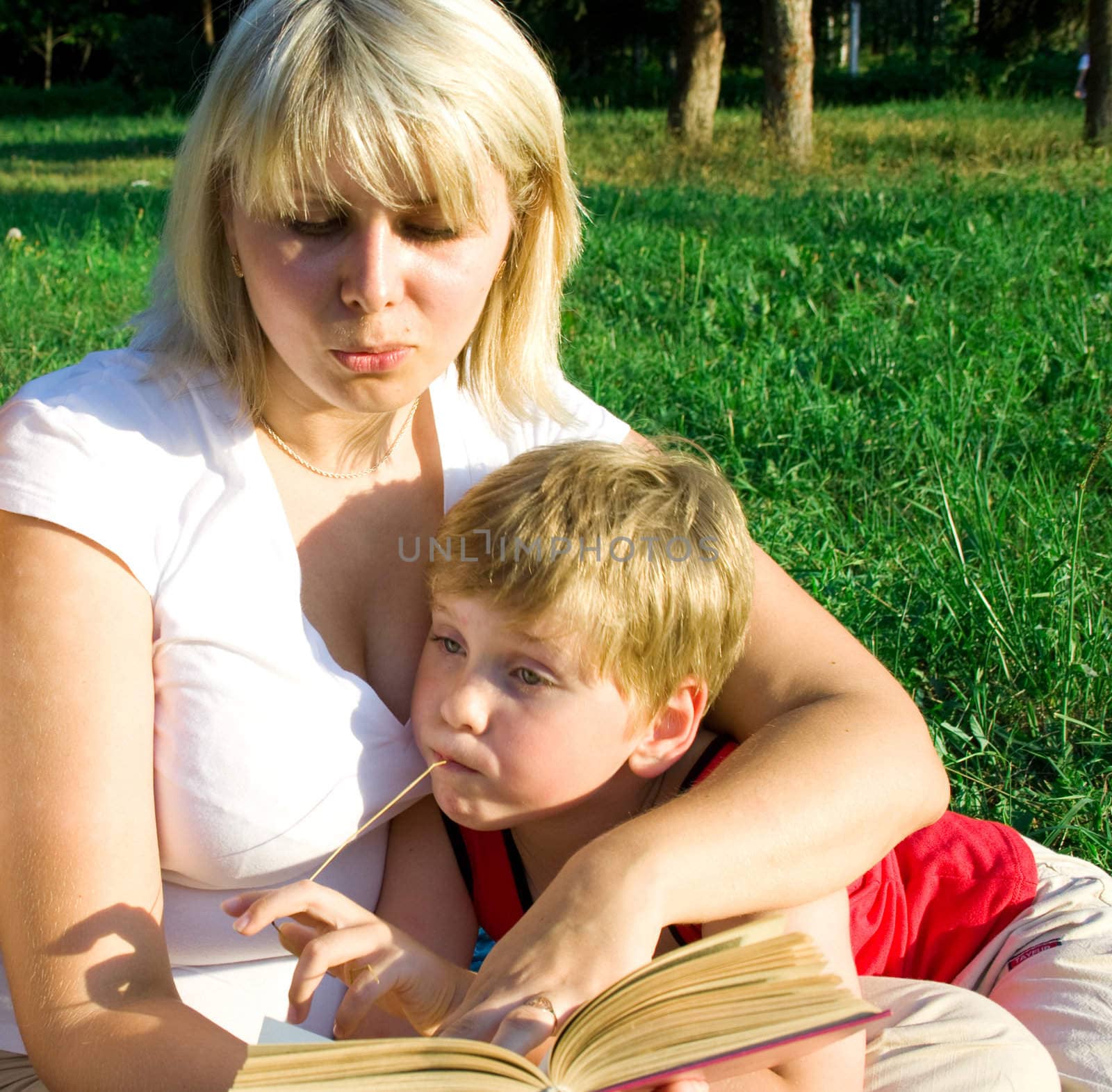 Mother sits on a grass with the son in a lap and reads to the boy the book